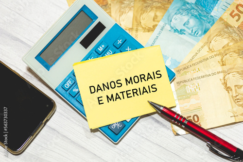 As palavras Danos morais e materiais escrita em um pedaço de papel que está sobre uma calculadora. Notas do Real Brasileiro na composição. Economia brasileira. photo