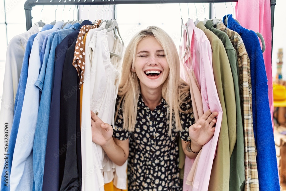 Young blonde woman searching clothes on clothing rack smiling and laughing hard out loud because funny crazy joke.