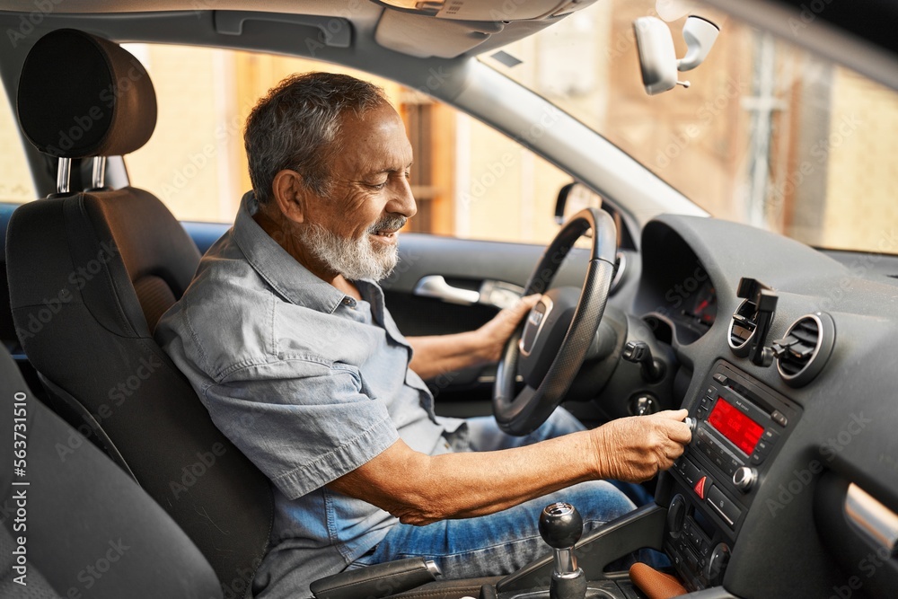 Senior grey-haired man sitting on car turning on radio at street