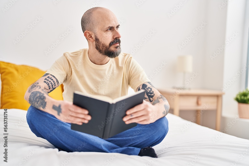 Young bald man reading book sitting on bed at bedroom Young bald man reading book sitting on bed at bedroom
