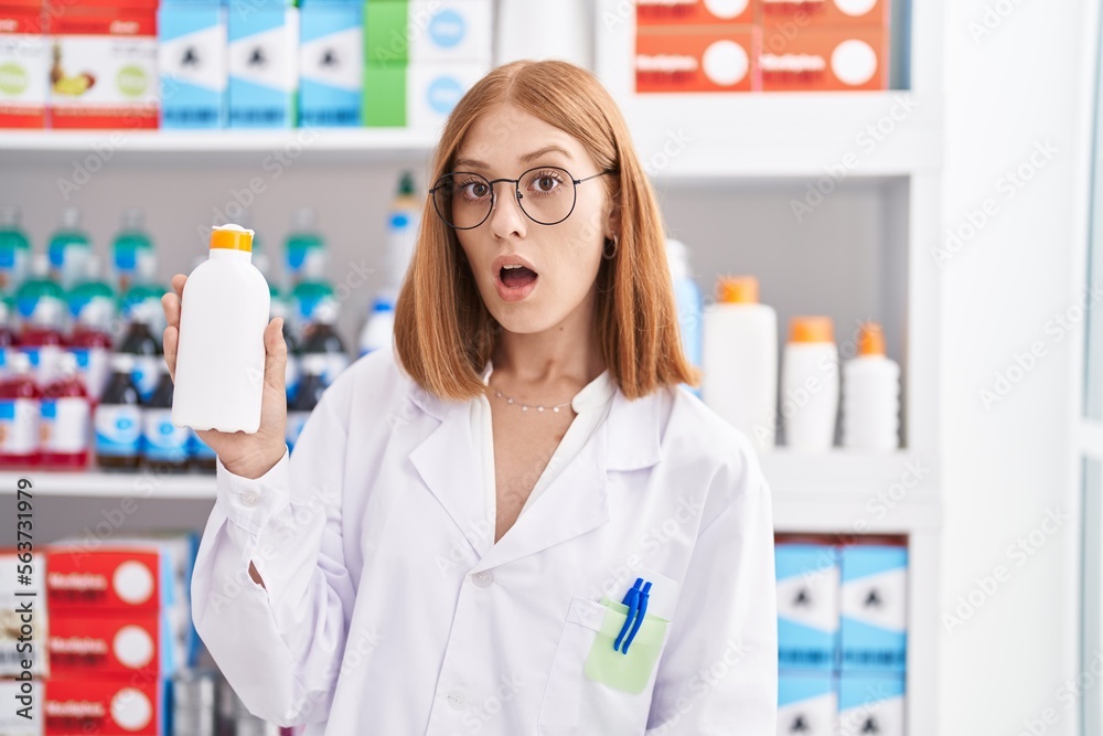 Young redhead woman working at pharmacy drugstore holding sun screen scared and amazed with open mouth for surprise, disbelief face