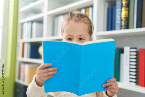 Adorable blonde girl student reading book standing at library school