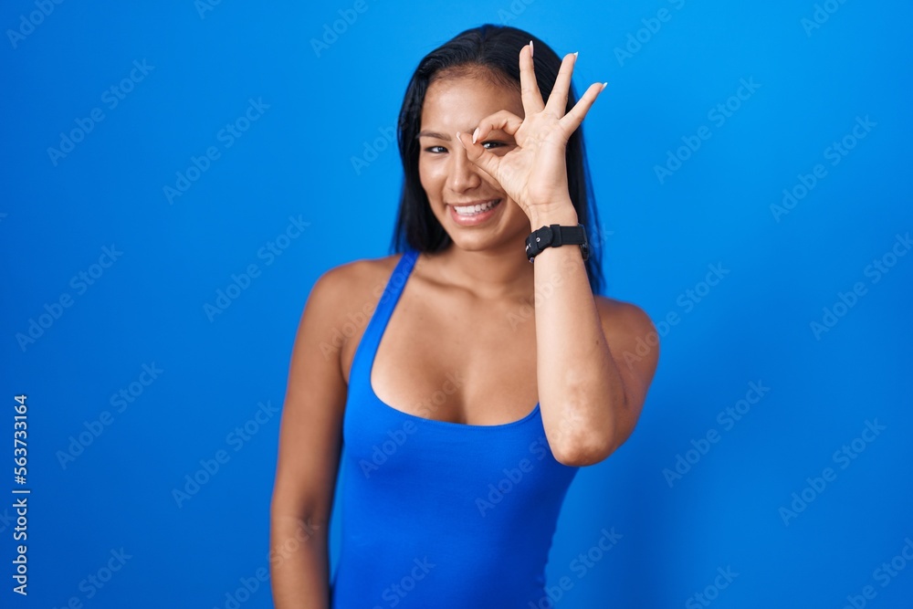 Hispanic woman standing over blue background doing ok gesture with hand smiling, eye looking through fingers with happy face. Hispanic woman standing over blue background doing ok gesture with hand smiling, eye looking through fingers with happy face.