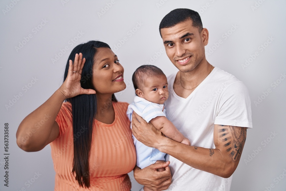 Young hispanic couple with baby standing together over isolated background smiling with hand over ear listening an hearing to rumor or gossip. deafness concept. Young hispanic couple with baby standing together over isolated background smiling with hand over ear listening an hearing to rumor or gossip. deafness concept.