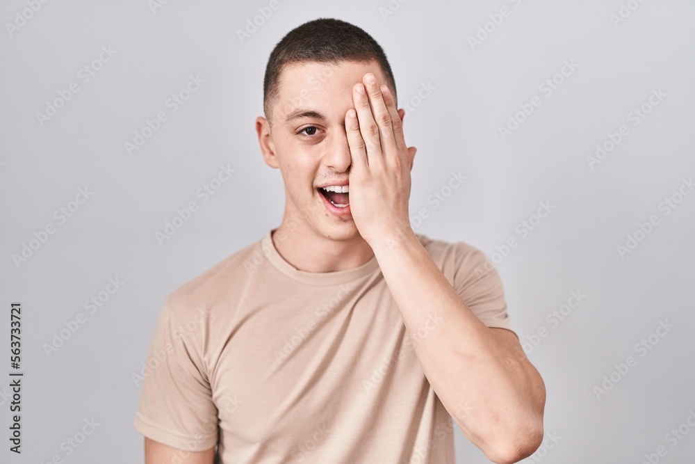 Young man standing over isolated background covering one eye with hand, confident smile on face and surprise emotion. Young man standing over isolated background covering one eye with hand, confident smile on face and surprise emotion.