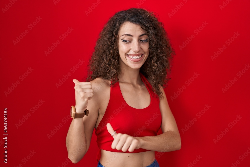 Hispanic woman with curly hair standing over red background pointing to the back behind with hand and thumbs up, smiling confident Hispanic woman with curly hair standing over red background pointing to the back behind with hand and thumbs up, smiling confident