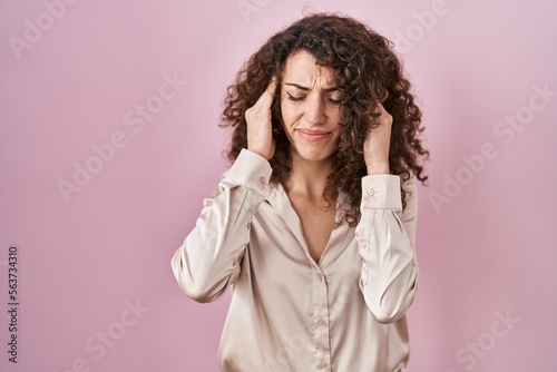 Hispanic woman with curly hair standing over pink background with hand on head, headache because stress. suffering migraine.