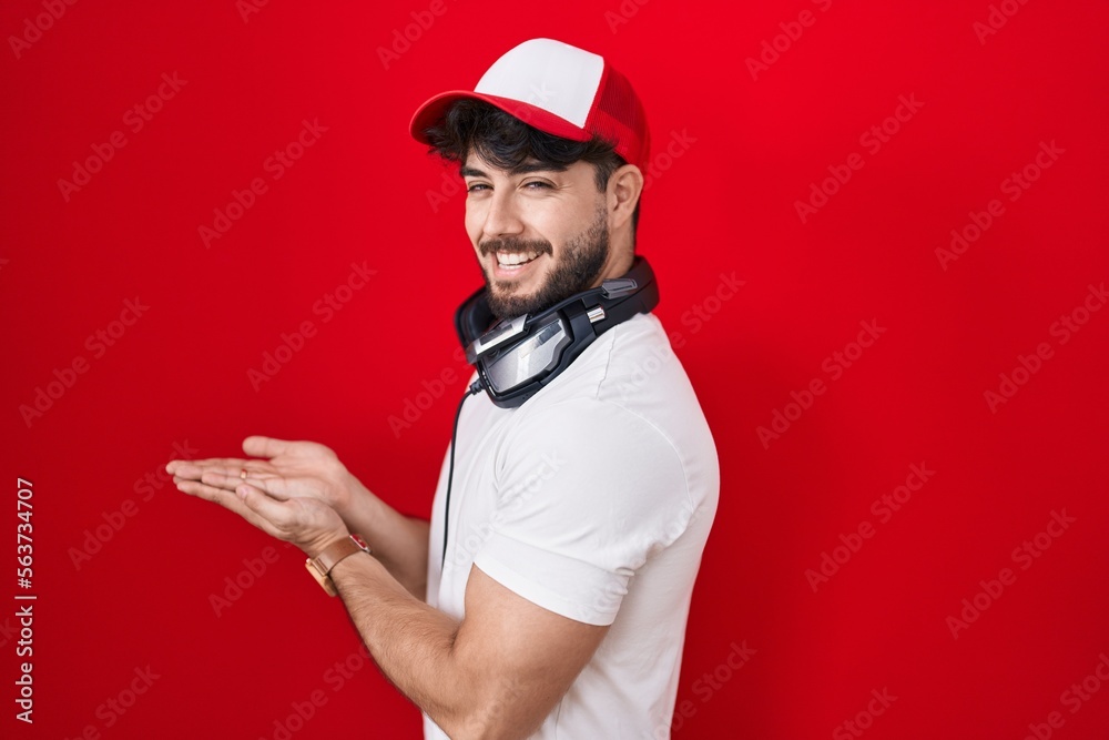 Hispanic man with beard wearing gamer hat and headphones pointing aside with hands open palms showing copy space, presenting advertisement smiling excited happy Hispanic man with beard wearing gamer hat and headphones pointing aside with hands open palms showing copy space, presenting advertisement smiling excited happy