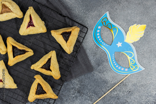 Jewish holiday Purim celebration concept. Traditional hamantasch or hamantaschen Haman's ears cookies, carnival mask, spin gragger (ratchet) noisemaker, and party decor on gray background.