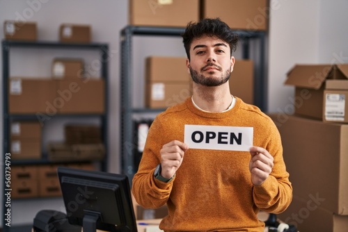 Hispanic man with beard working at small business ecommerce holding open sign relaxed with serious expression on face. simple and natural looking at the camera.