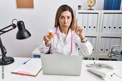 Young hispanic woman wearing doctor uniform holding pills at the clinic pointing up looking sad and upset, indicating direction with fingers, unhappy and depressed.