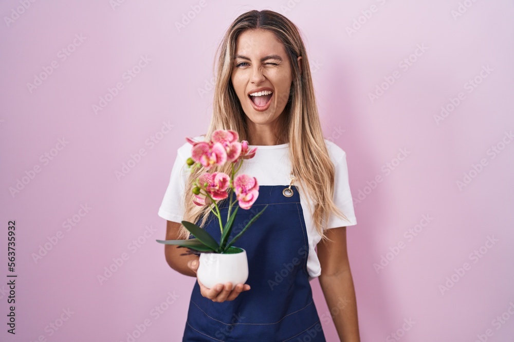 Young blonde woman wearing gardener apron holding plant winking looking at the camera with sexy expression, cheerful and happy face. Young blonde woman wearing gardener apron holding plant winking looking at the camera with sexy expression, cheerful and happy face.