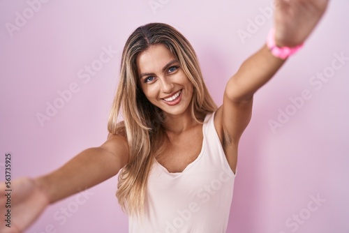 Young blonde woman standing over pink background looking at the camera smiling with open arms for hug. cheerful expression embracing happiness.
