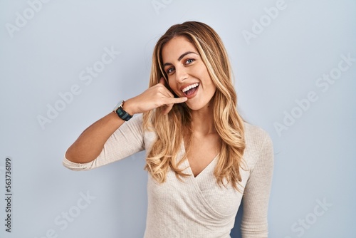 Young blonde woman standing over isolated background smiling doing phone gesture with hand and fingers like talking on the telephone. communicating concepts.