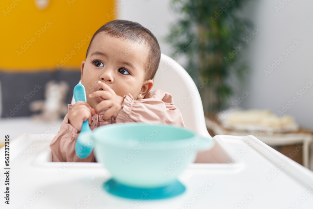 Adorable hispanic baby sitting on highchair holding spoon at bedroom Adorable hispanic baby sitting on highchair holding spoon at bedroom