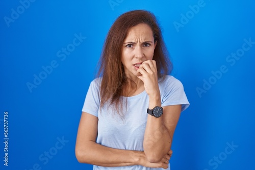Brunette woman standing over blue background looking stressed and nervous with hands on mouth biting nails. anxiety problem.