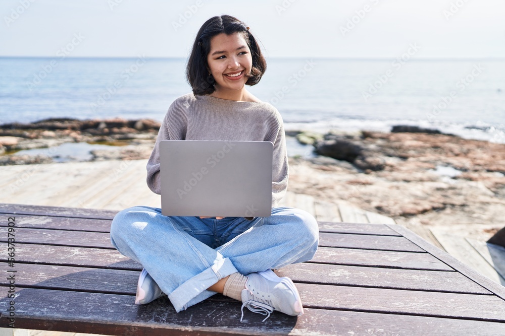 Young woman using laptop sitting on bench at seaside