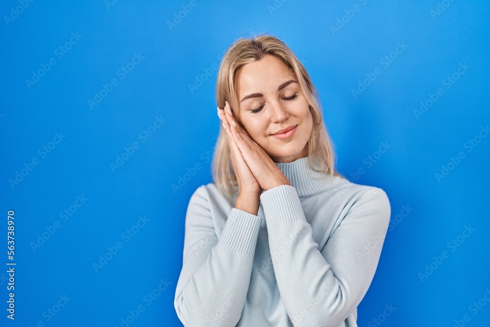 Young caucasian woman standing over blue background sleeping tired dreaming and posing with hands together while smiling with closed eyes. Young caucasian woman standing over blue background sleeping tired dreaming and posing with hands together while smiling with closed eyes.