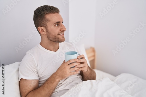 Young caucasian man drinking cup of coffee sitting on bed at bedroom