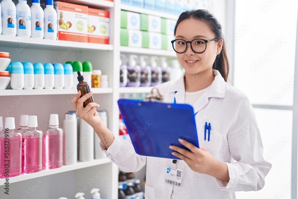 Young chinese woman pharmacist reading clipboard holding bottle at pharmacy