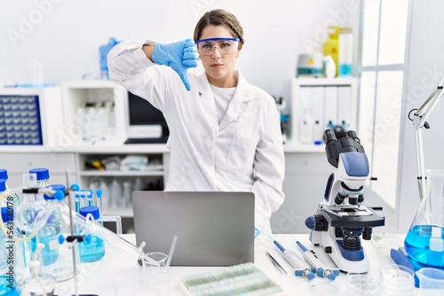 Young hispanic woman wearing scientist uniform working at laboratory with angry face  negative sign showing dislike with thumbs down  rejection concept
