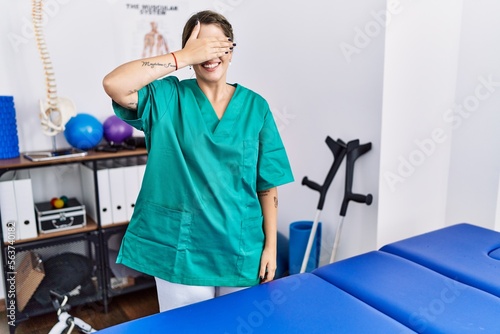 Young hispanic woman wearing physiotherapist uniform standing at clinic smiling and laughing with hand on face covering eyes for surprise. blind concept.
