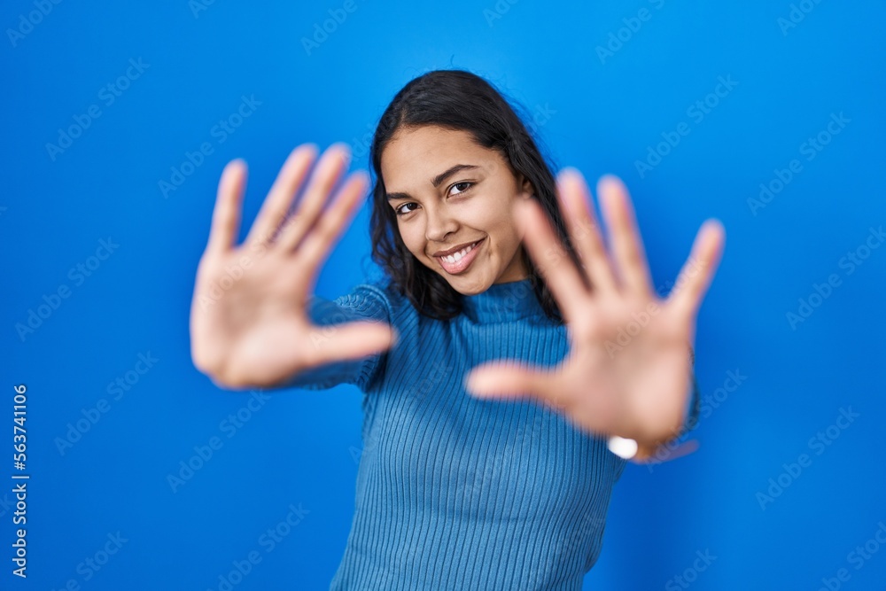 Young brazilian woman standing over blue isolated background doing frame using hands palms and fingers, camera perspective