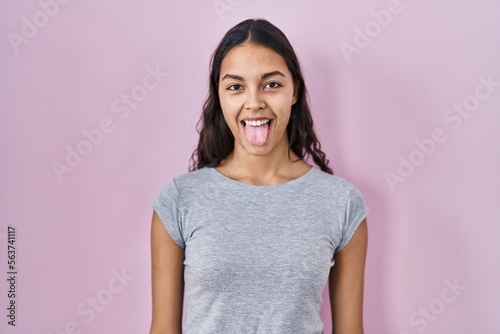 Young brazilian woman wearing casual t shirt over pink background sticking tongue out happy with funny expression. emotion concept.