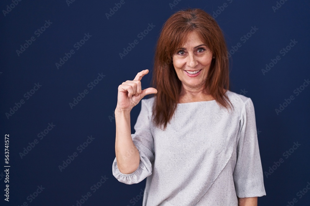 Middle age woman standing over blue background smiling and confident gesturing with hand doing small size sign with fingers looking and the camera. measure concept. Middle age woman standing over blue background smiling and confident gesturing with hand doing small size sign with fingers looking and the camera. measure concept.