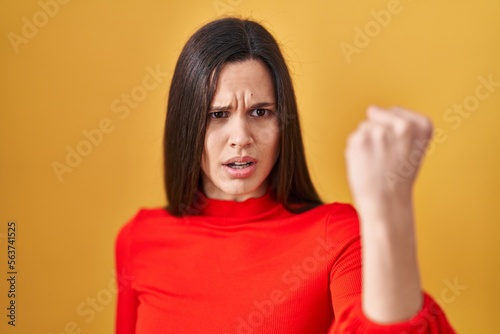 Young hispanic woman standing over yellow background angry and mad raising fist frustrated and furious while shouting with anger. rage and aggressive concept. © Krakenimages.com
