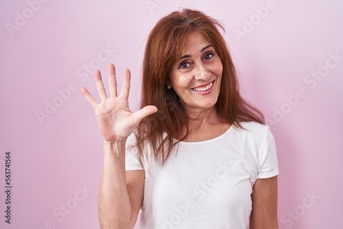 Middle age woman standing over pink background showing and pointing up with fingers number five while smiling confident and happy.