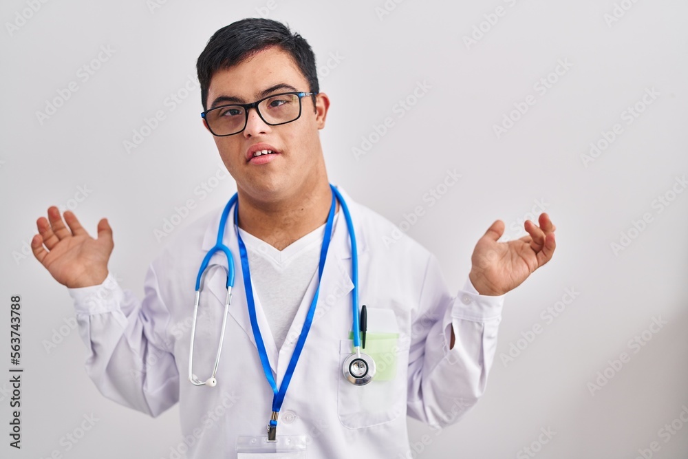 Young hispanic man with down syndrome wearing doctor uniform and stethoscope clueless and confused expression with arms and hands raised. doubt concept. Young hispanic man with down syndrome wearing doctor uniform and stethoscope clueless and confused expression with arms and hands raised. doubt concept.