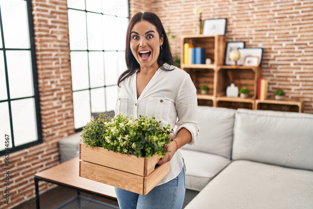 Young hispanic woman holding green plants at the living room celebrating crazy and amazed for success with open eyes screaming excited. Young hispanic woman holding green plants at the living room celebrating crazy and amazed for success with open eyes screaming excited.