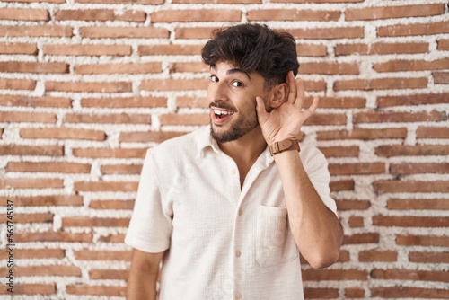 Arab man with beard standing over bricks wall background smiling with hand over ear listening an hearing to rumor or gossip. deafness concept.