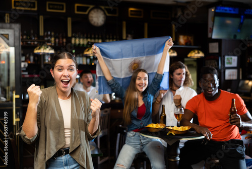 Happy young girl emotionally gesturing while watching football match in sports bar  cheering for favorite team of Argentina and celebrating victory