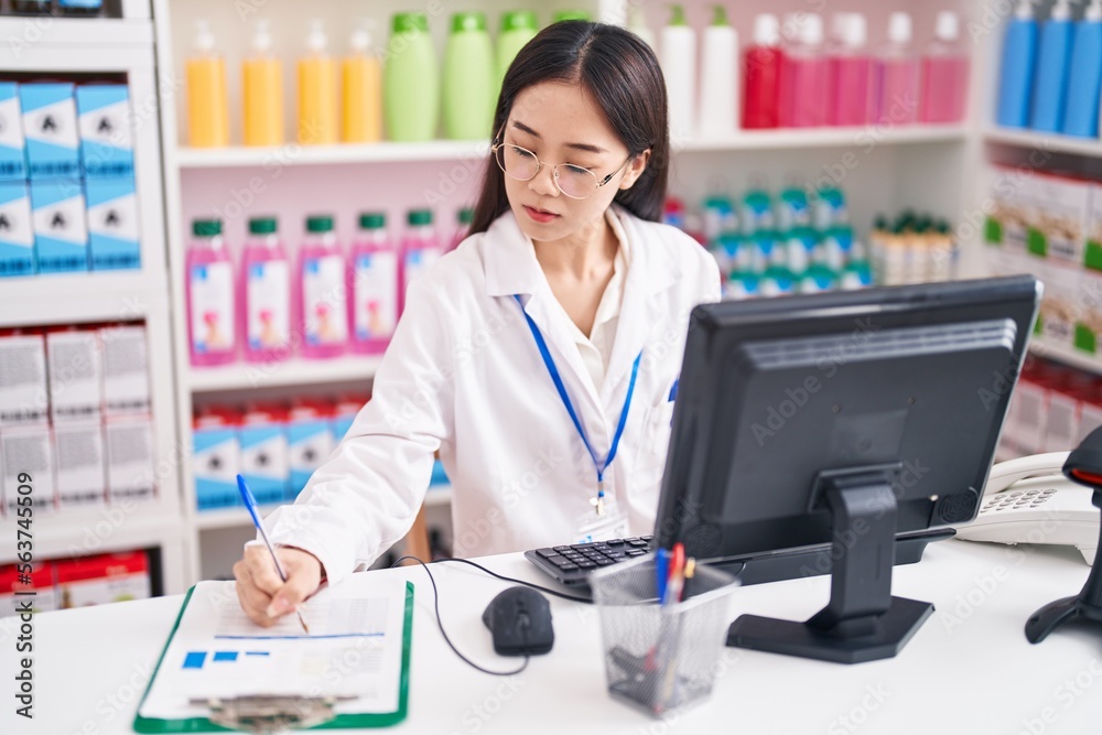 Young chinese woman pharmacist writing on document using computer at pharmacy Young chinese woman pharmacist writing on document using computer at pharmacy