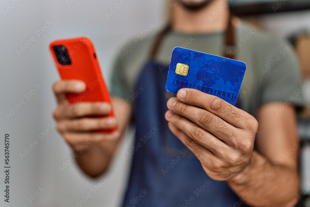 Fototapeta premium Young hispanic man business worker using smartphone and credit card at storehouse