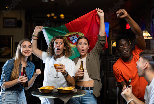 Group of Portugal football team fans spending time in bar, drinking bear and having fun. People with state flag in pub.