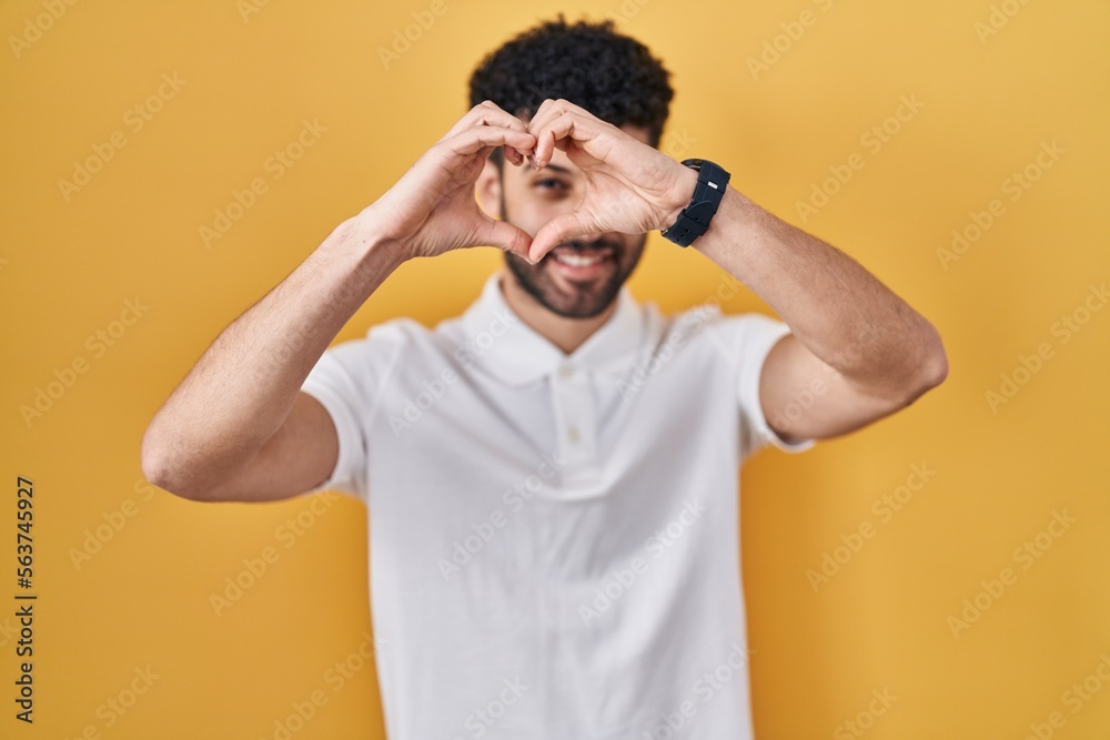Arab man standing over yellow background doing heart shape with hand and fingers smiling looking through sign Arab man standing over yellow background doing heart shape with hand and fingers smiling looking through sign