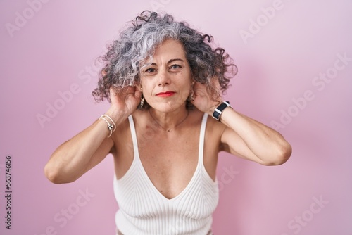 Middle age woman with grey hair standing over pink background trying to hear both hands on ear gesture, curious for gossip. hearing problem, deaf