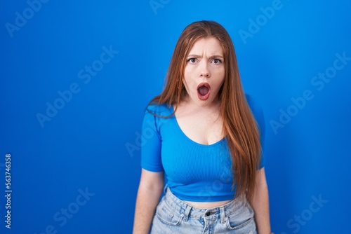 Redhead woman standing over blue background afraid and shocked with surprise and amazed expression, fear and excited face.