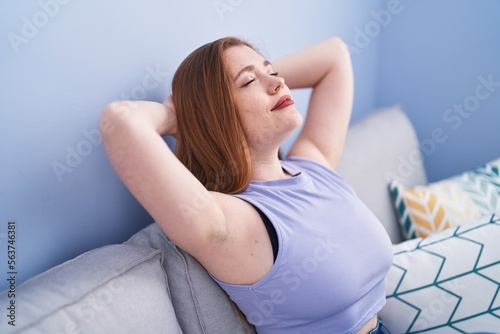 Young redhead woman relaxed with hands on head sitting on sofa at home
