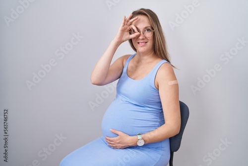 Young pregnant woman wearing band aid for vaccine injection smiling happy doing ok sign with hand on eye looking through fingers