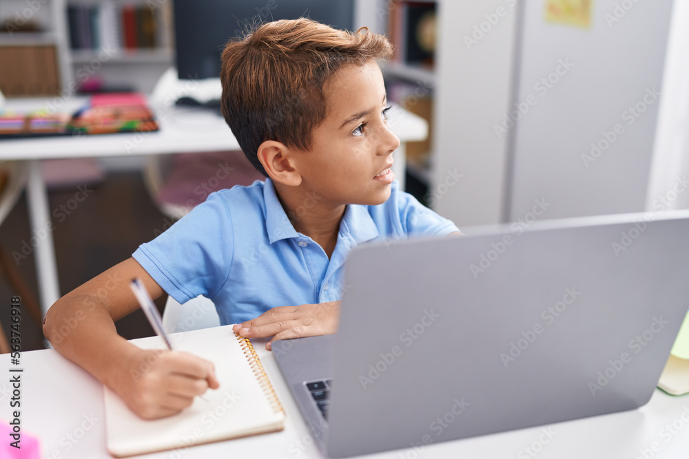 Adorable hispanic boy student using computer writing on notebook at classroom Adorable hispanic boy student using computer writing on notebook at classroom