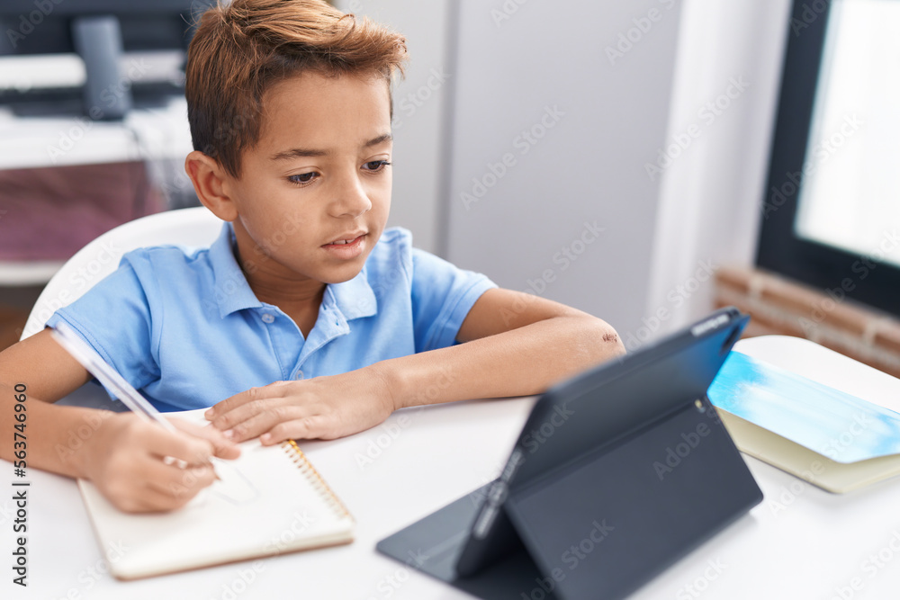 Adorable hispanic boy student sitting on table doing homework at classroom Adorable hispanic boy student sitting on table doing homework at classroom