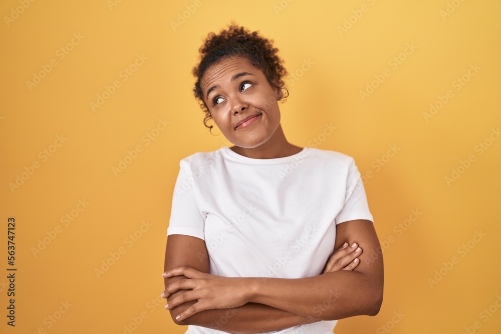 Young hispanic woman with curly hair standing over yellow background looking to the side with arms crossed convinced and confident Young hispanic woman with curly hair standing over yellow background looking to the side with arms crossed convinced and confident