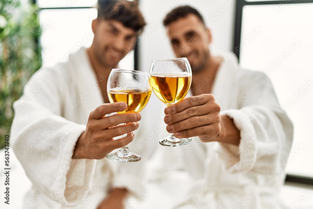 Two hispanic men couple toasting with champagne sitting on massage table at beauty center Two hispanic men couple toasting with champagne sitting on massage table at beauty center