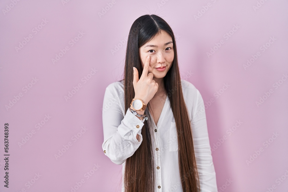 Chinese young woman standing over pink background pointing to the eye watching you gesture, suspicious expression Chinese young woman standing over pink background pointing to the eye watching you gesture, suspicious expression
