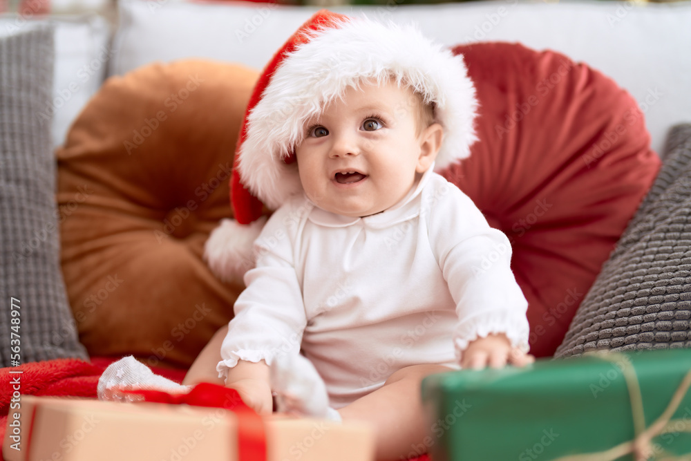 Adorable toddler wearing christmas hat sitting on sofa with present at home Adorable toddler wearing christmas hat sitting on sofa with present at home