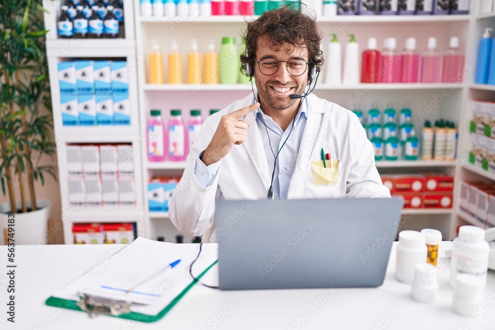 Hispanic young man working at pharmacy drugstore working with laptop smiling happy pointing with hand and finger